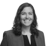A woman with wavy dark hair, wearing a blazer and a blouse, smiles at the camera against a plain white background.