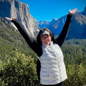 allison-knight Woman in a white vest stands with arms raised, smiling in front of a scenic mountain landscape with clear blue sky.