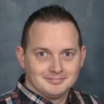 A man with short brown hair and blue eyes is wearing a plaid shirt, looking at the camera with a neutral expression against a gray background.