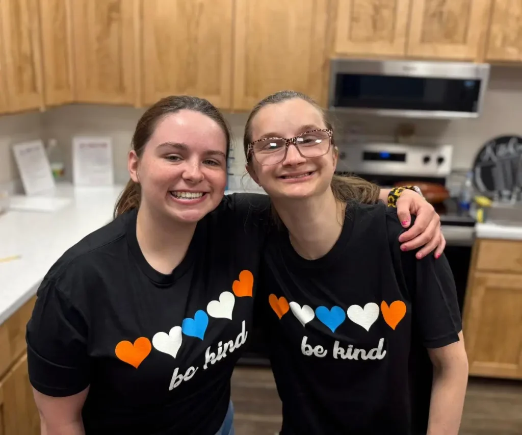 Two people stand in a kitchen, smiling with arms around each other. Both wear matching black 
