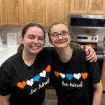 Two people stand in a kitchen, smiling with arms around each other. Both wear matching black "be kind" shirts with blue, orange, and white hearts.