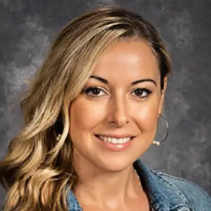 dulce-s Woman with long blonde hair, wearing hoop earrings and a denim jacket, smiling at the camera in front of a gray mottled background.