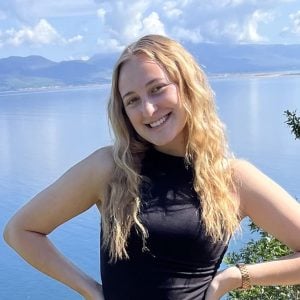 anna-s A woman with long blonde hair in a black sleeveless top stands smiling in front of a lake with mountains and cloudy sky in the background.