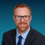 A man with red hair, beard, and glasses is wearing a suit, blue shirt, and tie, posing for a professional headshot against a plain blue background.